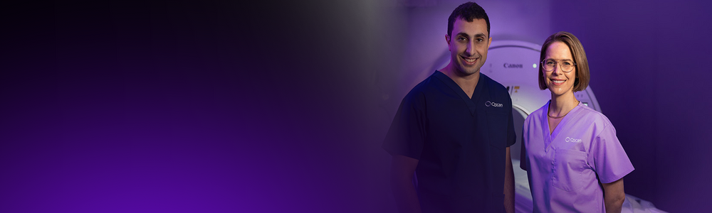 Two Qscan radiology staff members in scrubs standing together in front of a scanner, smiling at the camera.