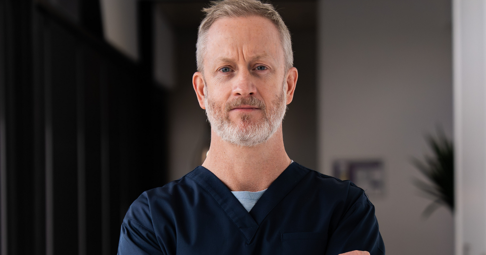 Dr Brian Carey, a Qscan radiologist, standing in a clinic hallway in navy scrubs, looking directly at the camera.