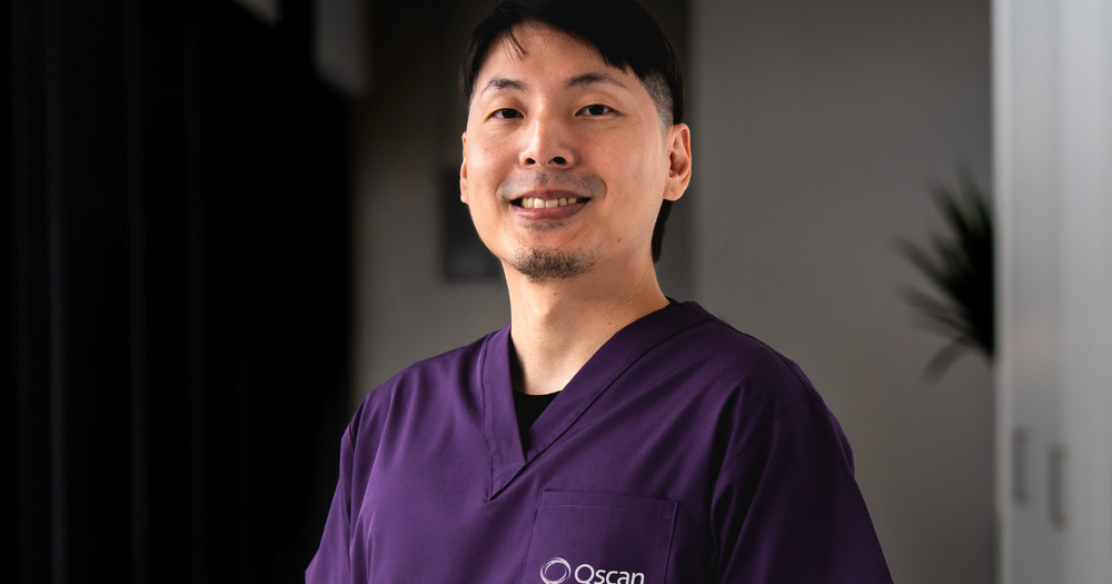 Dr Randy, a Qscan radiologist, standing in a clinic hallway in purple scrubs, looking directly at the camera.