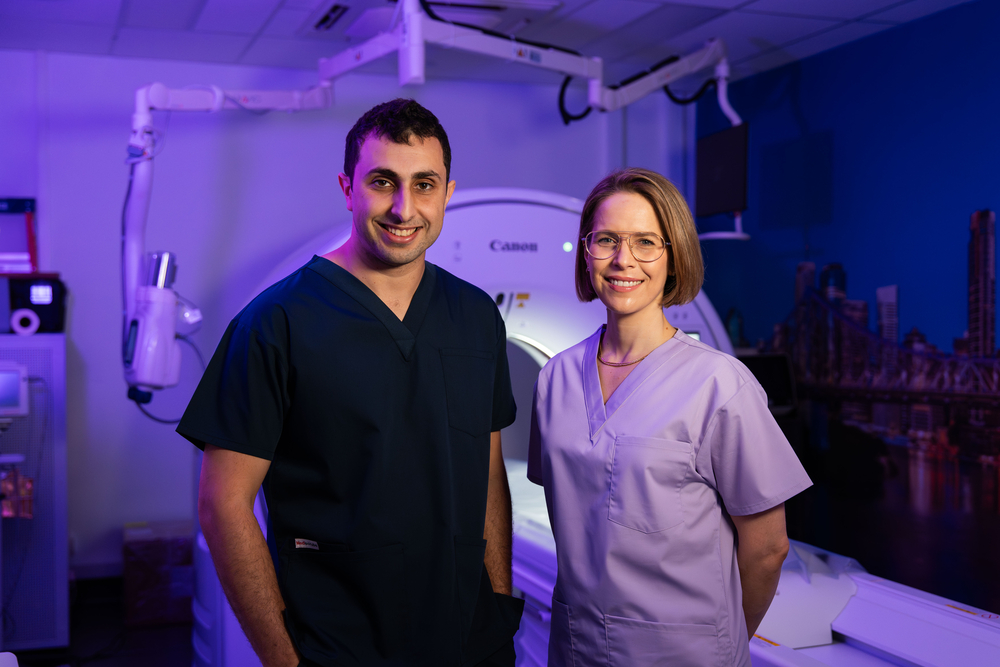 Two Qscan radiology staff members in scrubs standing together in front of a scanner, smiling at the camera.