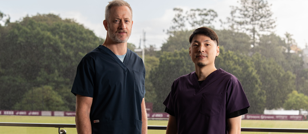 Dr Brian Carey and Dr Rany Huang, Qscan radiologists, standing together outdoors in scrubs with a green, tree-lined backdrop.
