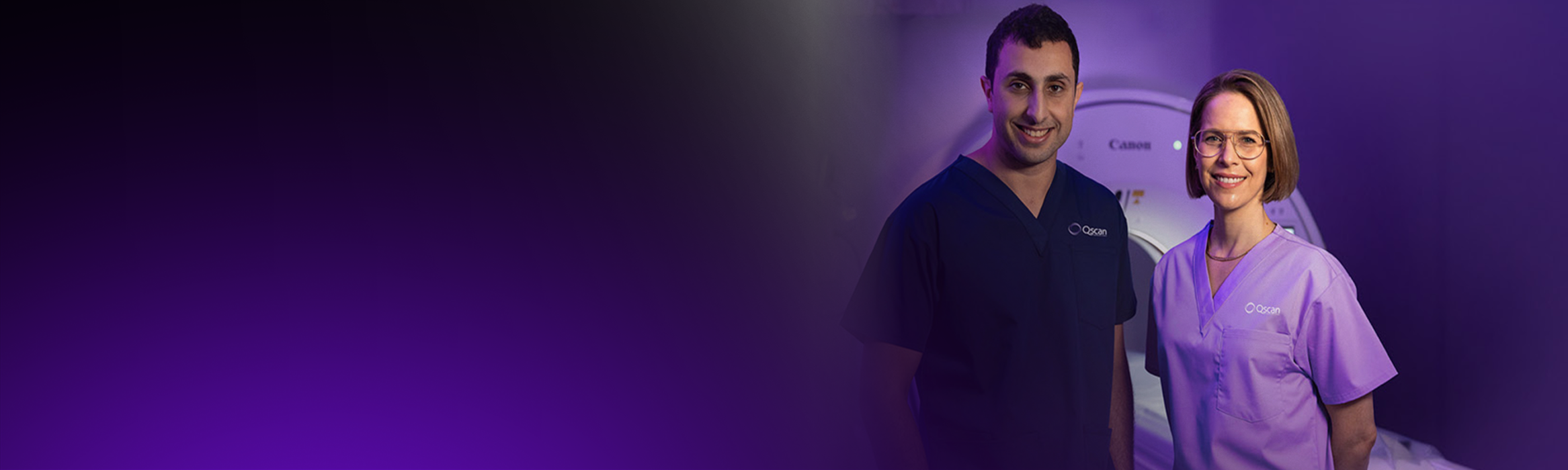 Two Qscan radiology staff members in scrubs standing together in front of a scanner, smiling at the camera.