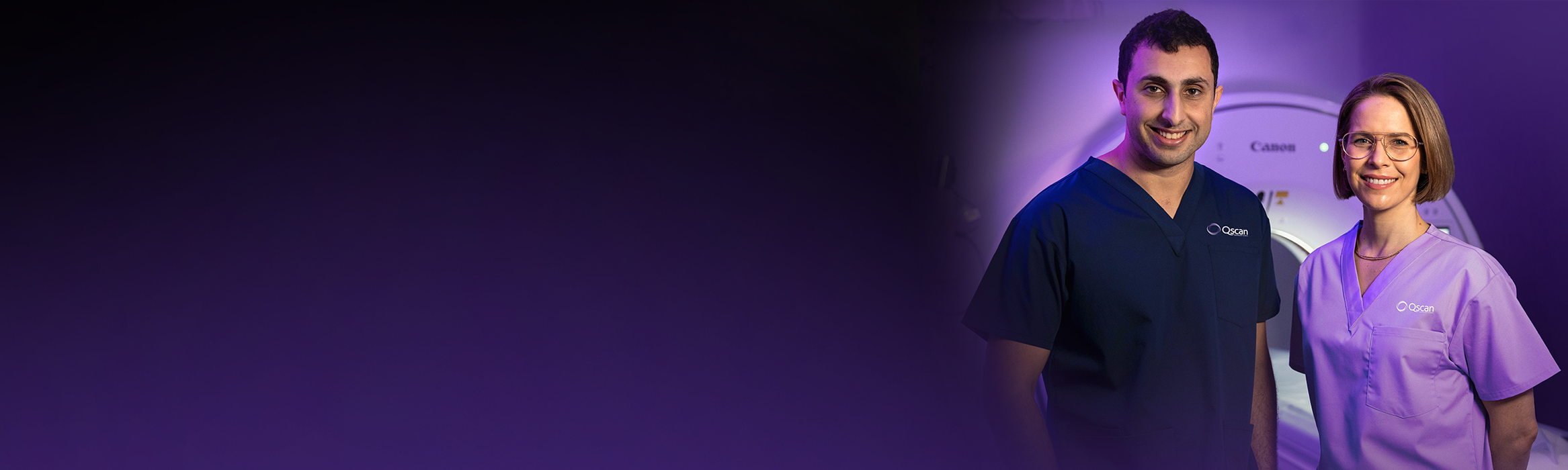 Two Qscan radiology staff members in scrubs standing together in front of a scanner, smiling at the camera.
